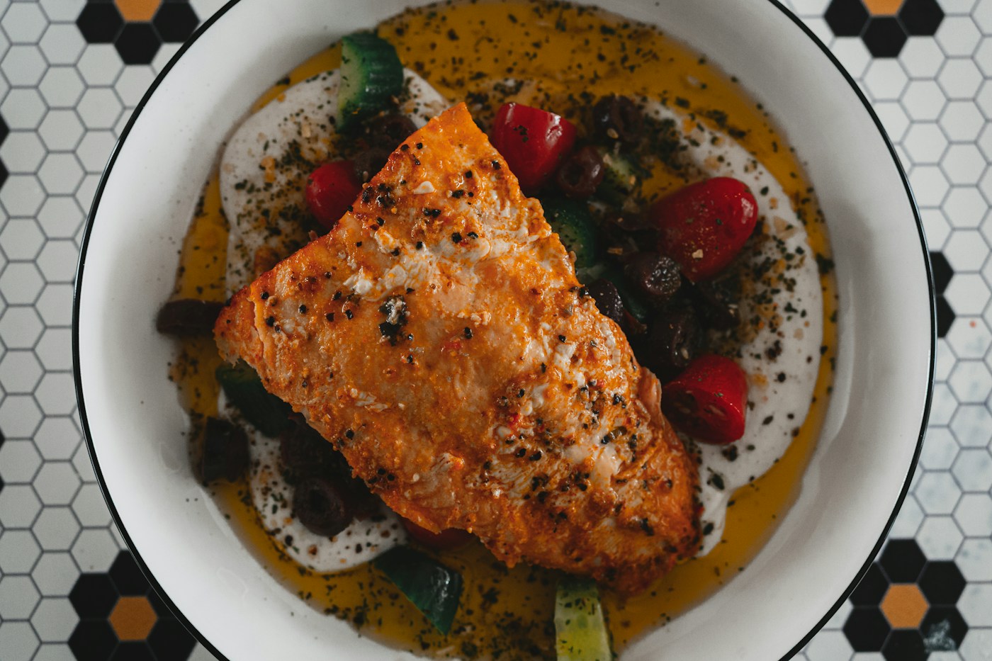Filete de pescado servido en un cuenco blanco con tomates cherry, aceitunas y verduras sobre mesa de mosaicos